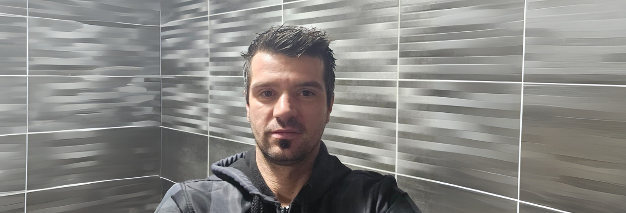 Muškarac u tamnoj jakni stoji ispred zida s modernim sivim keramičkim pločicama i gleda ravno u kameru.