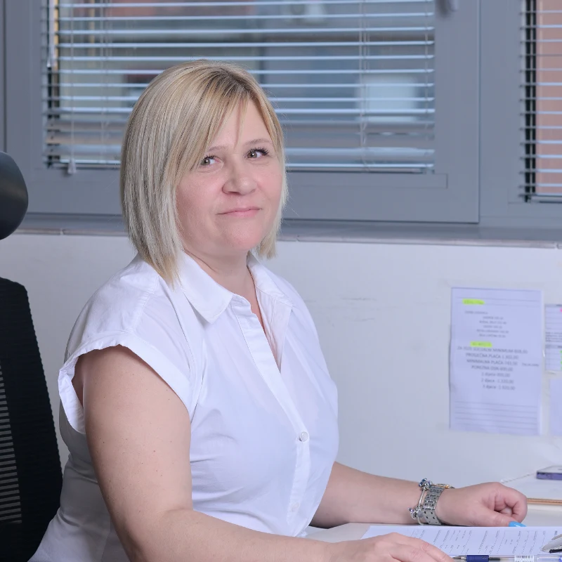 Žena s plavom kosom u bijeloj košulji sjedi za uredskim stolom, gleda u kameru i smiješi se.