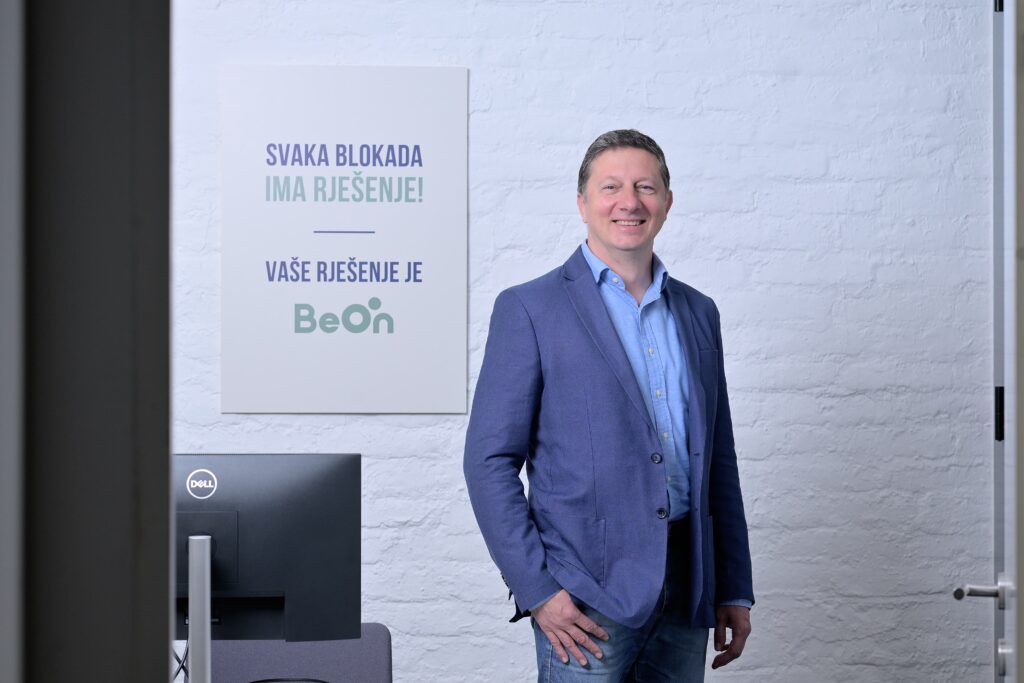 Muškarac u plavom sakou i svijetloplavoj košulji stoji nasmijan u uredu ispred zida s natpisom 'Svaka blokada ima rješenje! Vaše rješenje je BeOn'