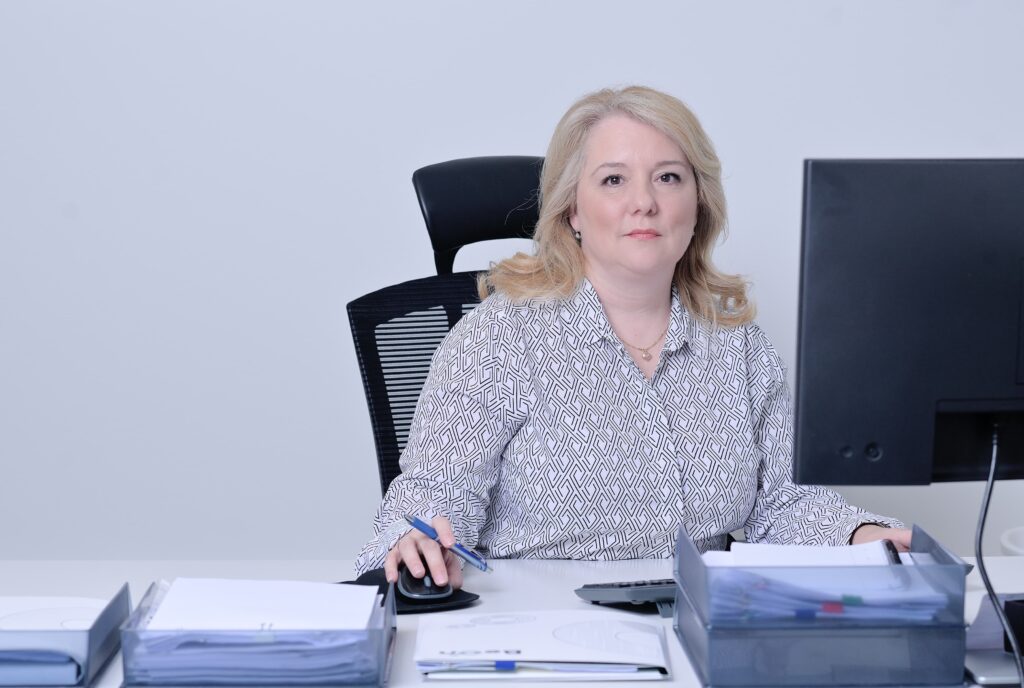 Žena sjedi za uredskim stolom ispred računala, drži olovku u ruci i gleda prema kameri. Na stolu su dokumenti i uredski pribor.