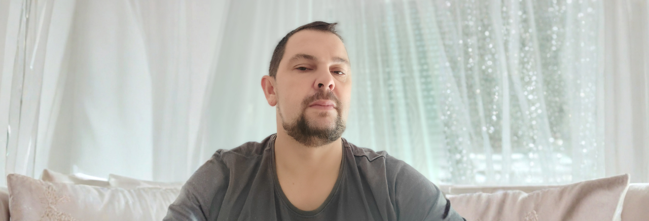 Muškarac kratke kose i brade sjedi na svijetloj sofi ispred prozora s bijelim zavjesama; nosi tamnosivu majicu i gleda ravno u kameru.