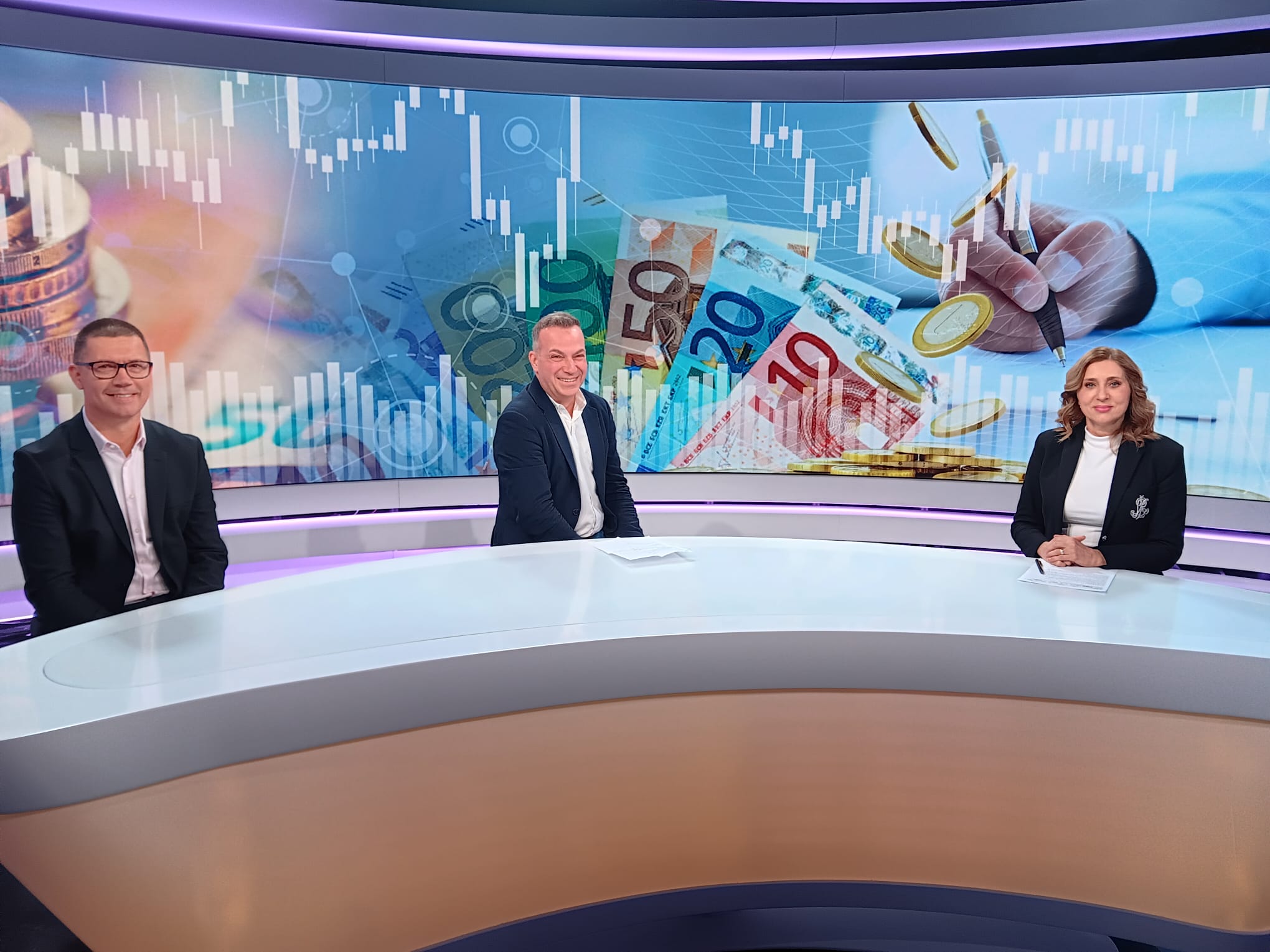Tri osobe sjede za studijskim stolom u modernom televizijskom studiju. Iza njih je veliki digitalni ekran s prikazom novčanica eura, kovanica i grafova koji simboliziraju financijska tržišta.