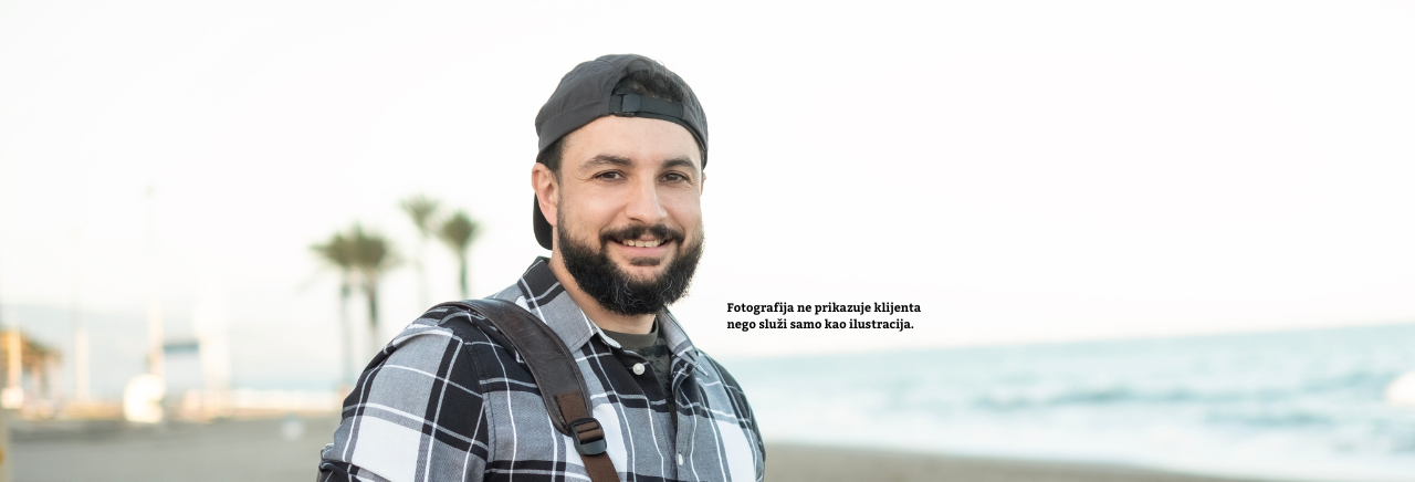 Mladić s bradom i kapom unatrag, u kariranoj košulji s remenom ruksaka preko ramena, nasmiješen stoji uz obalu mora.