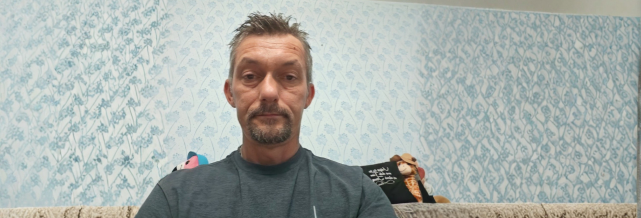 Muškarac sjedi na kauču i gleda u kameru, u pozadini je svijetloplava tapeta s uzorkom i nekoliko plišanih igračaka.