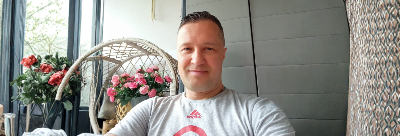 Nasmijani muškarac u sivoj majici sjedi u svijetlom interijeru pored stolice od ratana i buketa ružičastih ruža, u pozadini su veliki prozori.