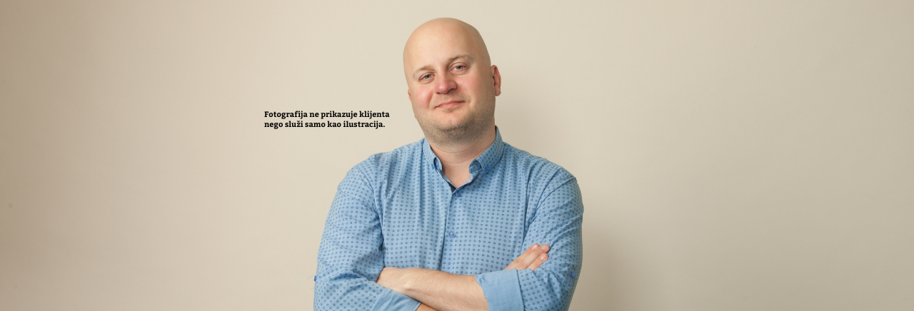 Muškarac s prekriženim rukama u plavoj košulji stoji na svijetloj neutralnoj pozadini; lijevo je tekst: “Fotografija ne prikazuje klijenta nego služi samo kao ilustracija.”