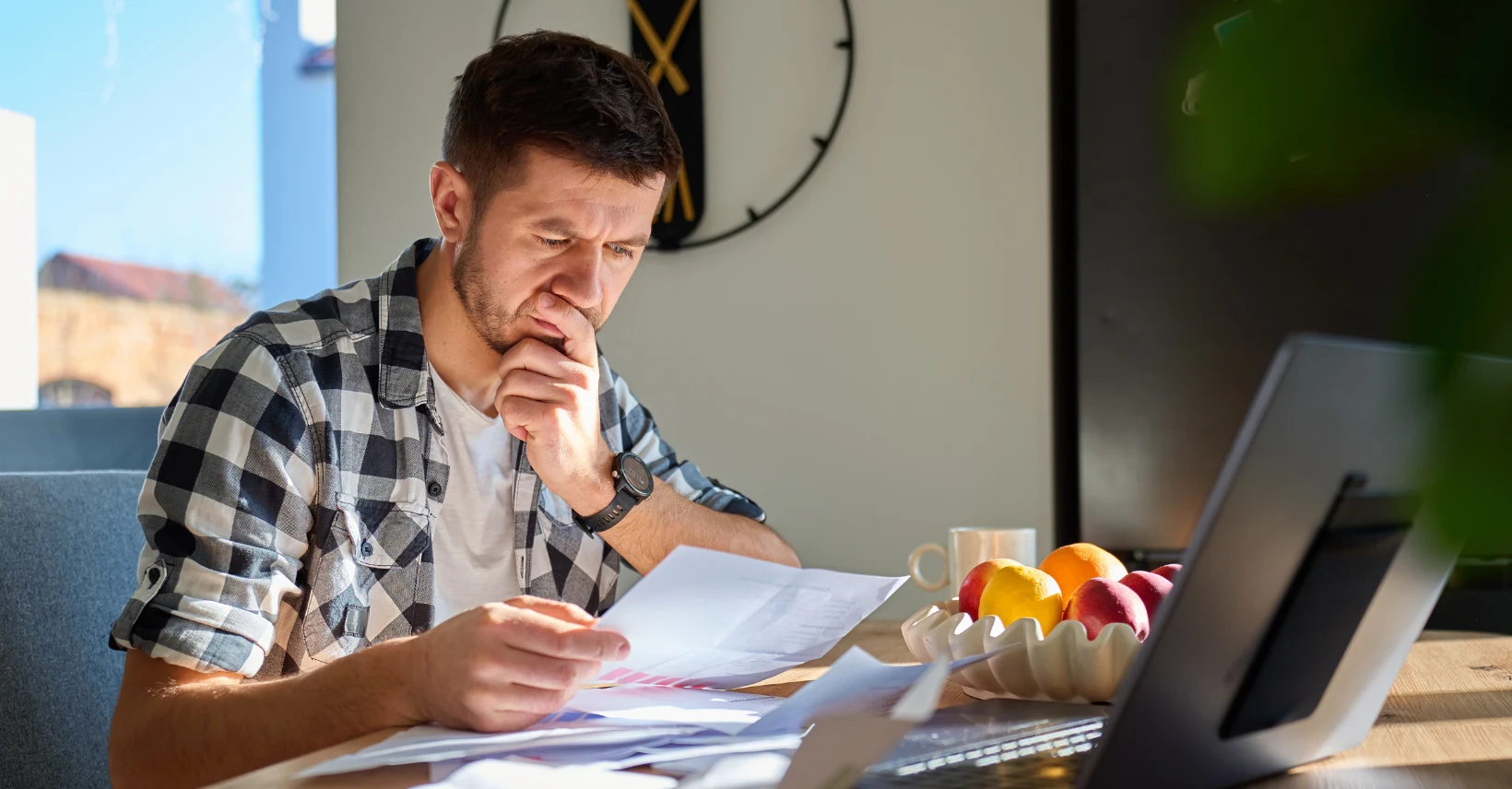 Muškarac pregledava račune i papire za stolom pokraj otvorenog laptopa.