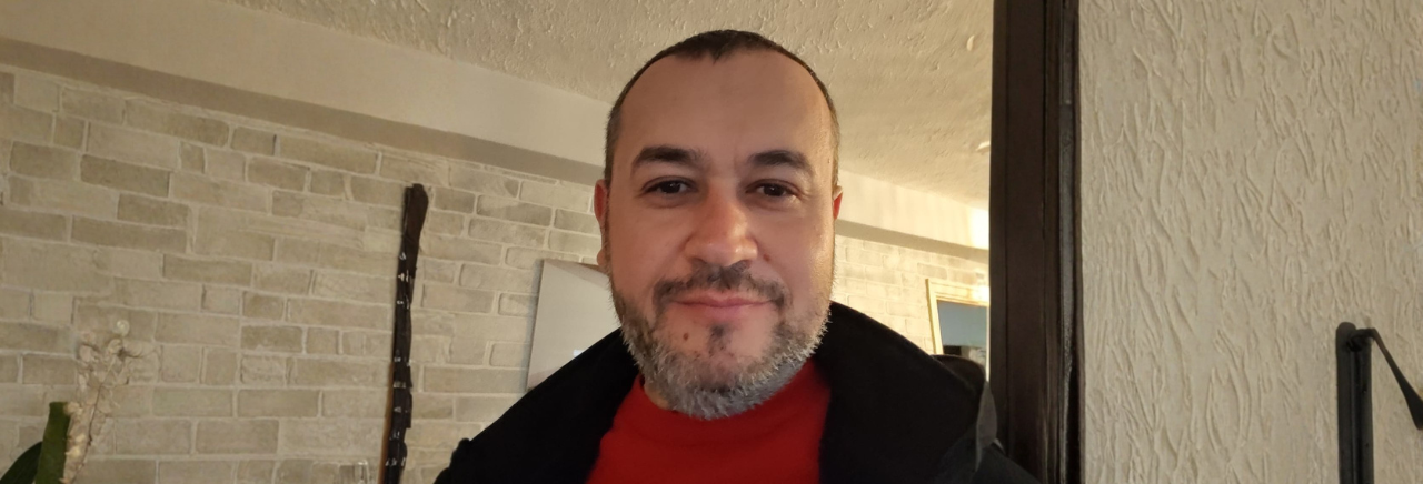 Muškarac snima selfie u zatvorenom prostoru, u crvenoj majici i tamnoj jakni; u pozadini su svijetli zid od ciglica i ogledalo.