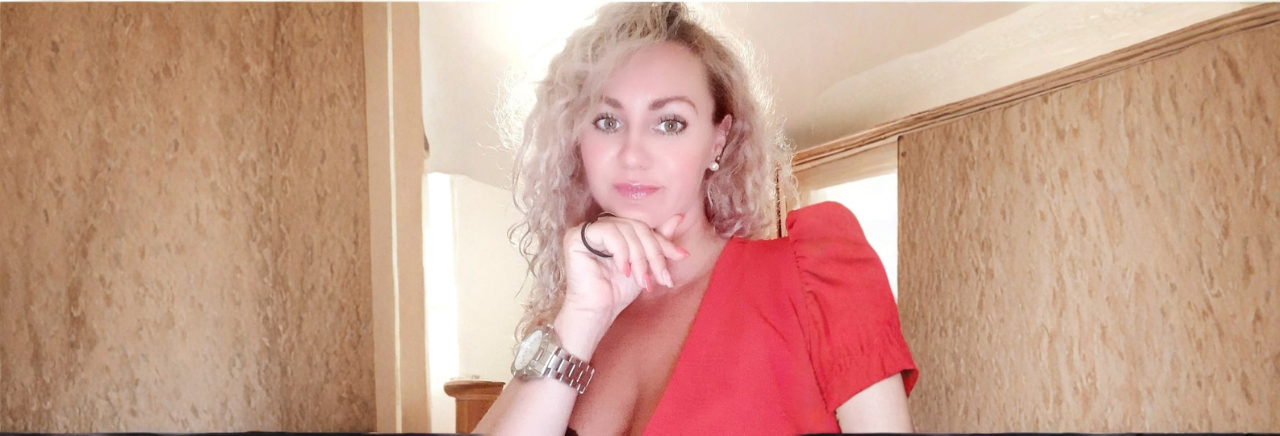 Žena svijetle kovrčave kose pozira naslonjena na ruku, odjevena u crvenu haljinu, u interijeru s toplim tonovima i prirodnim svjetlom.