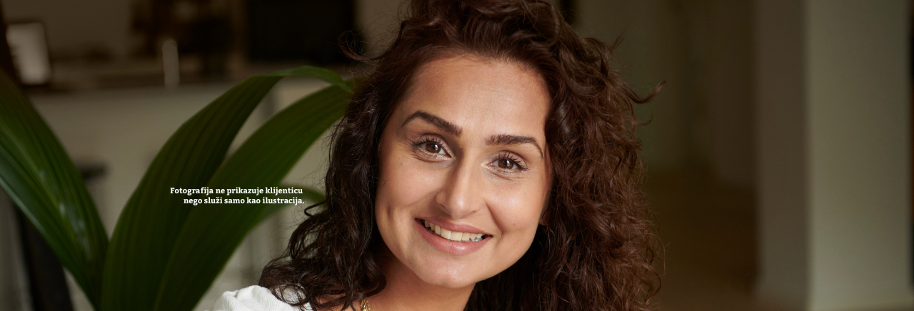 Portret nasmiješene žene s kovrčavom kosom u opuštenom interijeru, ilustrativna fotografija uz tekstualnu napomenu da osoba nije stvarna klijentica.