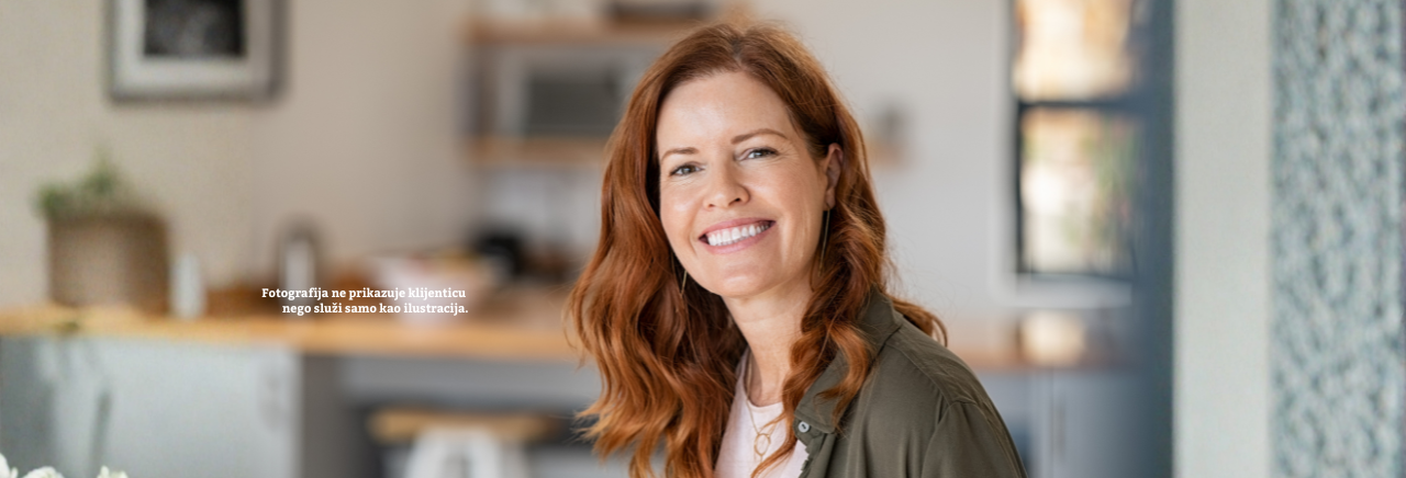 Žena srednjih godina s crvenom kosom nasmijano gleda u kameru, stoji u svijetlom, moderno uređenom interijeru kuhinje; fotografija ilustrativnog karaktera.